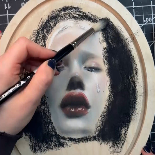 A hand uses a paintbrush to shade a portrait on an oval wooden panel, showing a pale face with dark hair, red lips, and a tear on one cheek.
